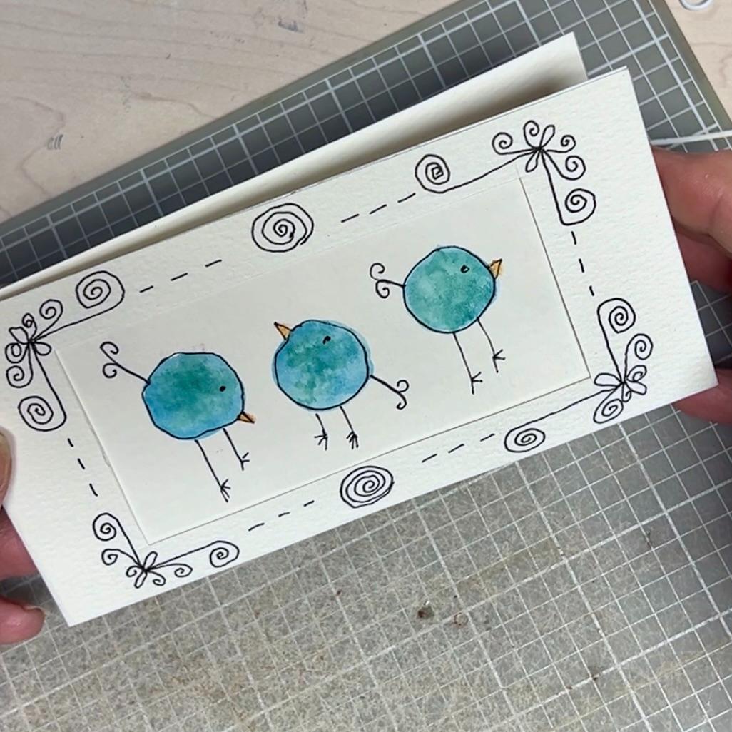 A handmade card features three blue, round birds drawn with watercolor and ink, surrounded by a whimsical black doodle border with swirls. The card is held above a gridded cutting mat.