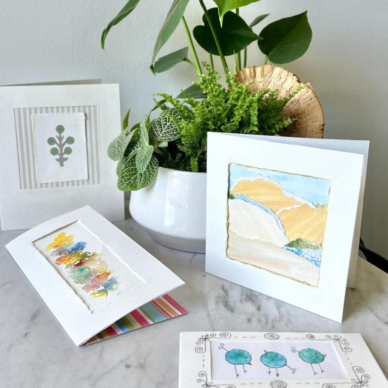 Four handmade greeting cards with nature-themed designs are displayed on a marble surface next to a white pot containing green plants. The cards feature artwork of leaves, abstract shapes, landscapes, and trees.