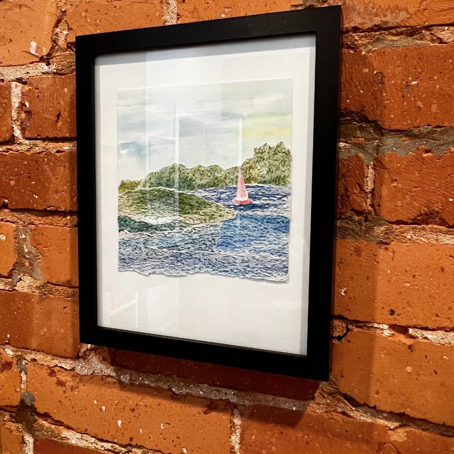 A framed watercolor painting of a pink sailboat on blue water with green islands in the background hangs on a brick wall.