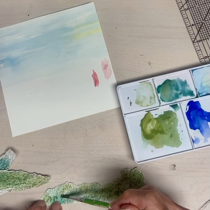 A person uses a green brush to paint a textured strip of paper. Nearby are a square palette with blue and green watercolor paints, a piece of watercolor paper, and a ruler on a light wooden surface.