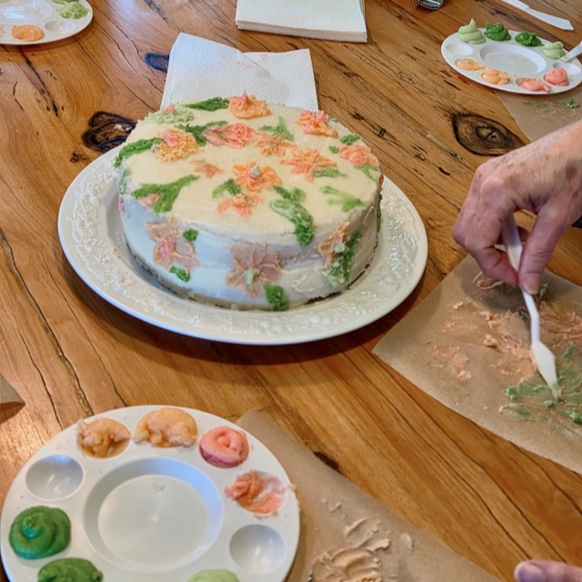 A person decorates a frosted cake with pastel-colored floral designs on a wooden table. Plates with dabs of colored icing and plastic forks are arranged nearby, along with napkins.