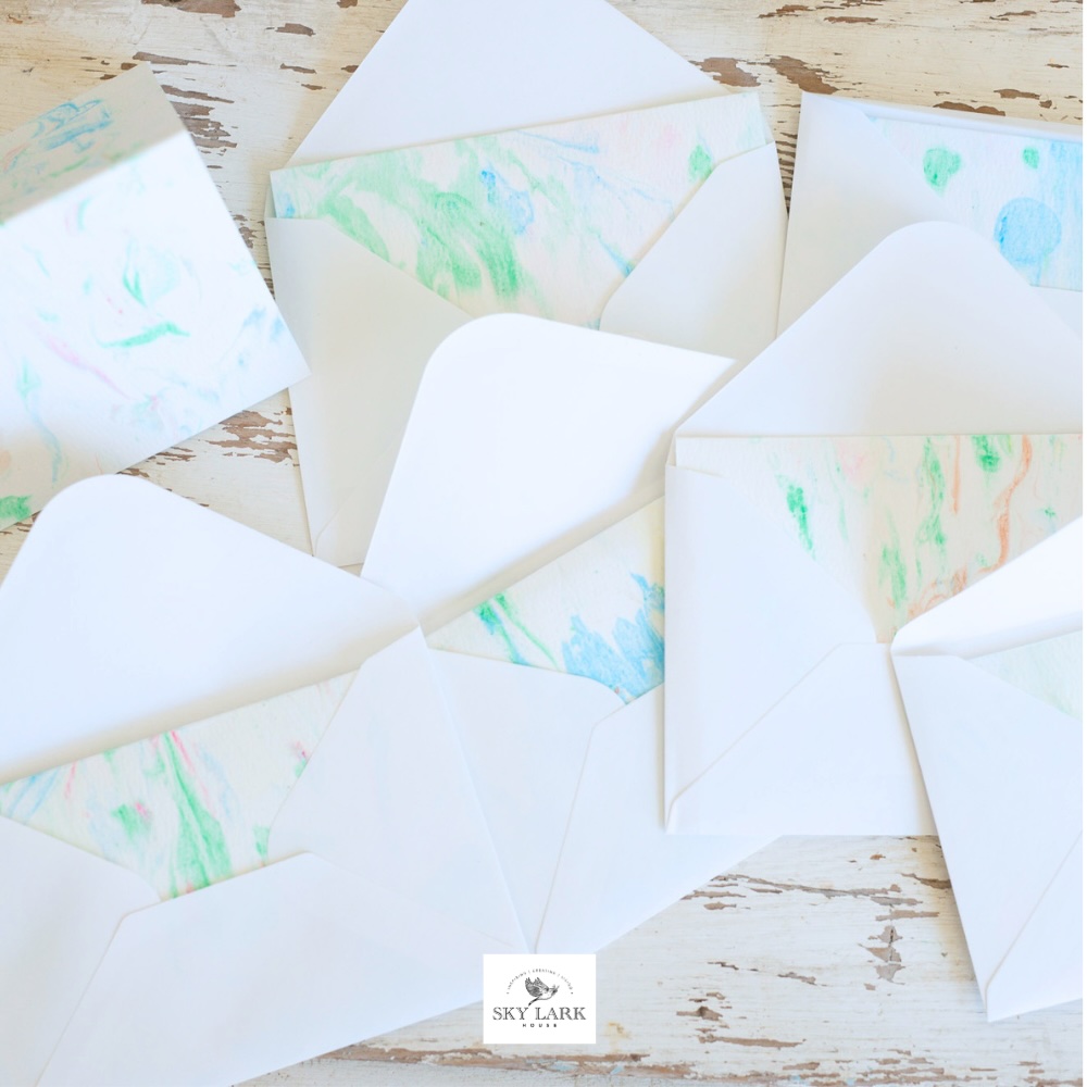 Several open white envelopes with pastel-colored, marbled liners are arranged on a rustic, white wooden surface. A faint Skylark logo appears near the center bottom of the image.