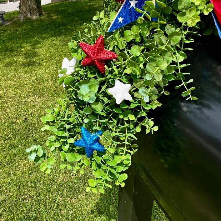 A DIY patriotic mailbox swag of greenery tops a black mailbox, accented with red, white, and blue glittery star ornaments for a festive touch. Grass and a tree trunk are visible in the background.