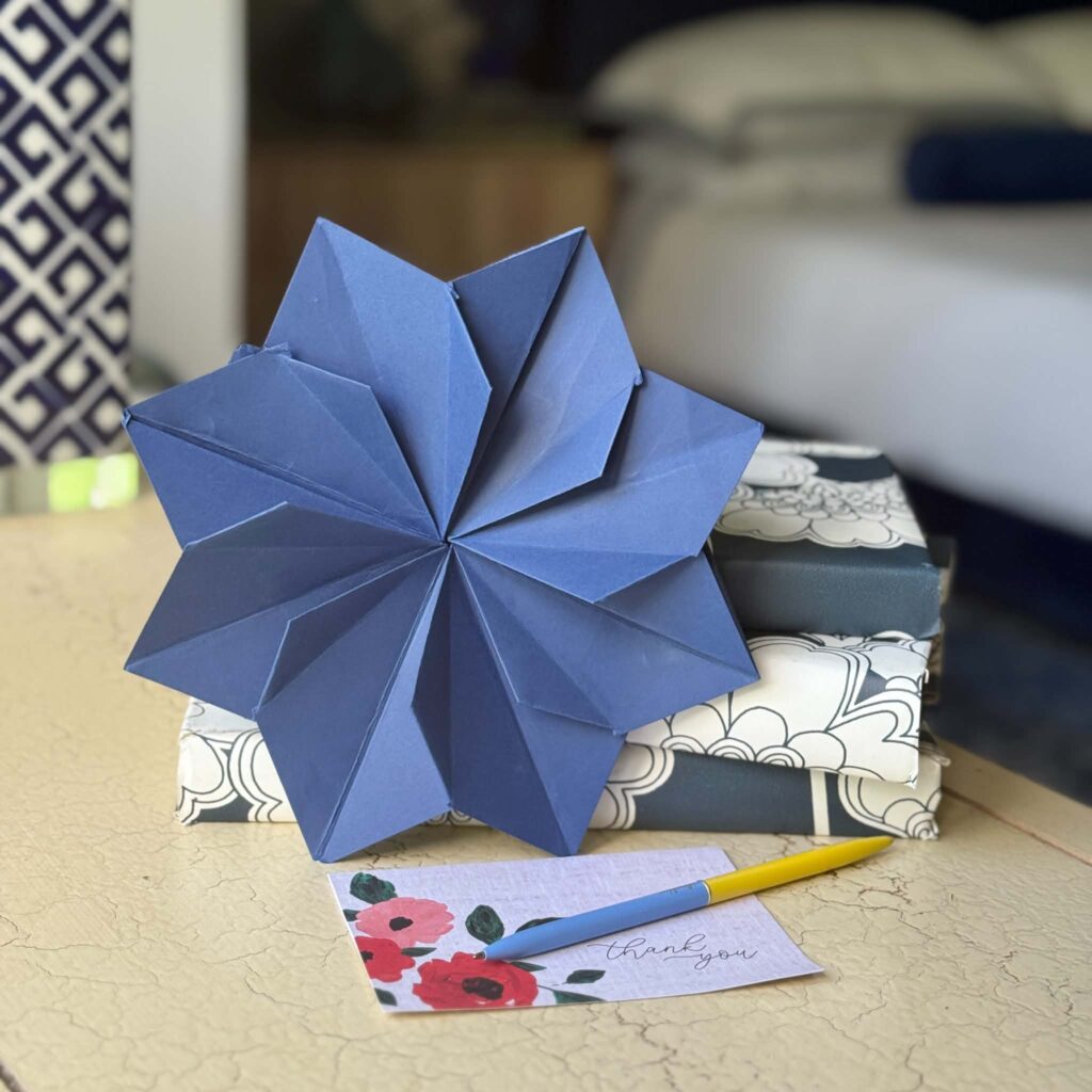 A large blue origami star rests on two stacked, patterned gift boxes. In front is a thank you card with painted flowers and a yellow pen, all arranged on a beige table.