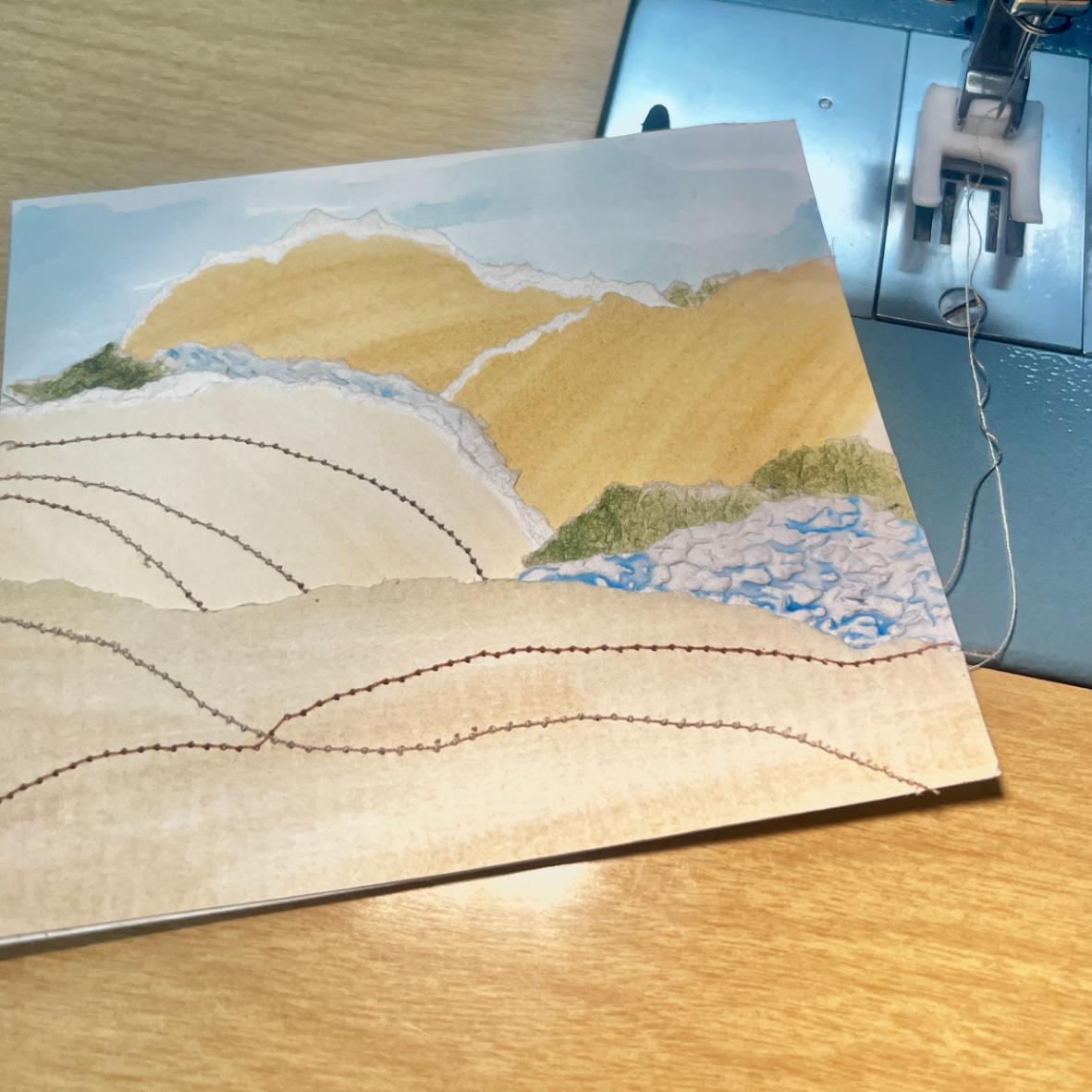 A landscape collage with layered paper and stitched thread lines is placed next to a sewing machine on a wooden surface. The artwork depicts hills, water, and greenery in soft, natural colors.