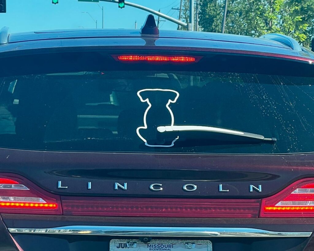A Lincoln SUV’s rear windshield wiper is shaped like a dog’s tail and is attached to a white dog silhouette decal on the rear window, making it look like the dog's tail wags when the wiper moves.