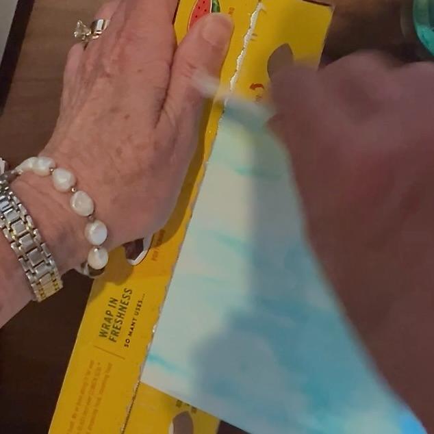 A person’s hands are pulling painted watercolor paper against the sharp serrated edge of a plastic wrap box to tear the paper