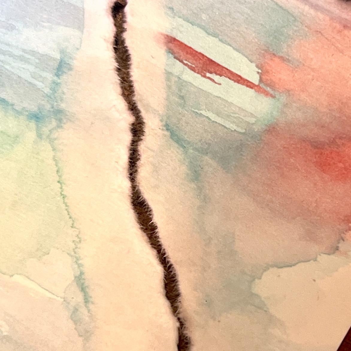 A close-up of torn watercolor paper with soft pastel hues of blue, red, and green, showing a jagged brown tear running vertically through the center of the image.