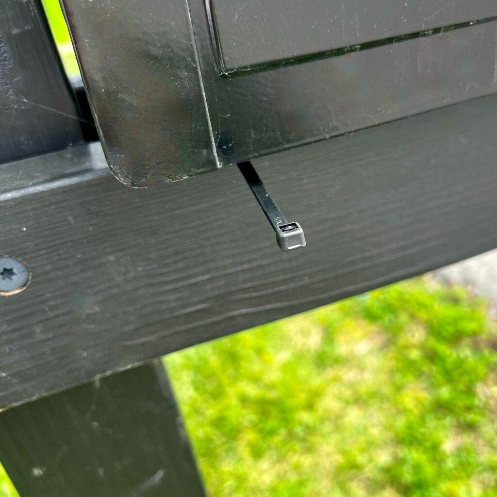 A close-up of a black zip tie securing two black wooden boards, perfect for assembling a DIY patriotic mailbox swag, with a grassy area visible in the blurred background.