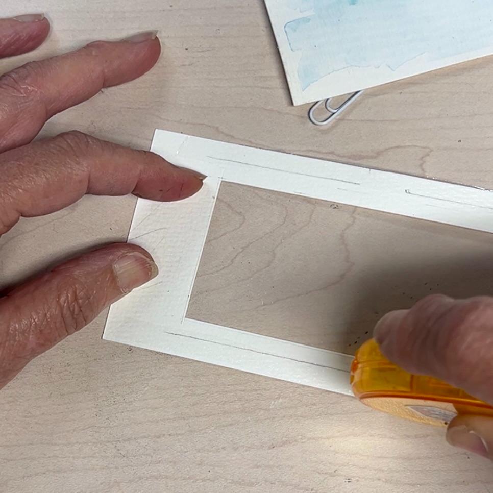 A person’s hands are holding a rectangular piece of white paper with a cut-out window while applying adhesive tape along the edge, on a light wooden surface. Another paper with a paperclip is partially visible above.