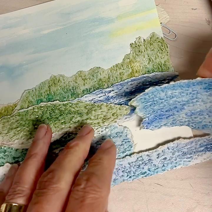 A person tears a sheet of textured paper with a painted landscape, featuring green hills, blue water, and a light sky, using their hands.