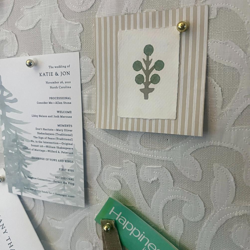 A close-up of a fabric pinboard holds a wedding invitation, a striped card with a simple green plant illustration, and a green card partially visible at the bottom, all held with gold thumbtacks.