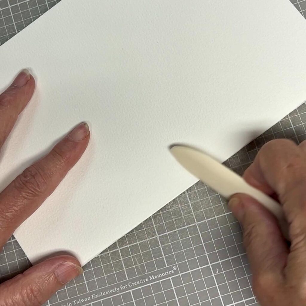A person uses a bone folder to crease a sheet of white textured paper placed on a gray grid cutting mat. Two hands are visible, holding the paper and the tool.
