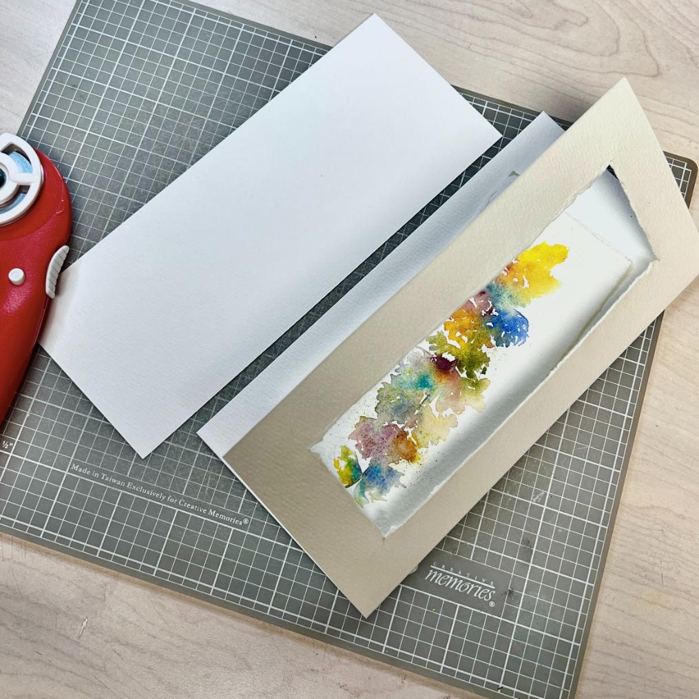 A handmade card with a watercolor design is being assembled on a cutting mat. The card features a colorful abstract pattern, and a red cutting tool lies nearby.