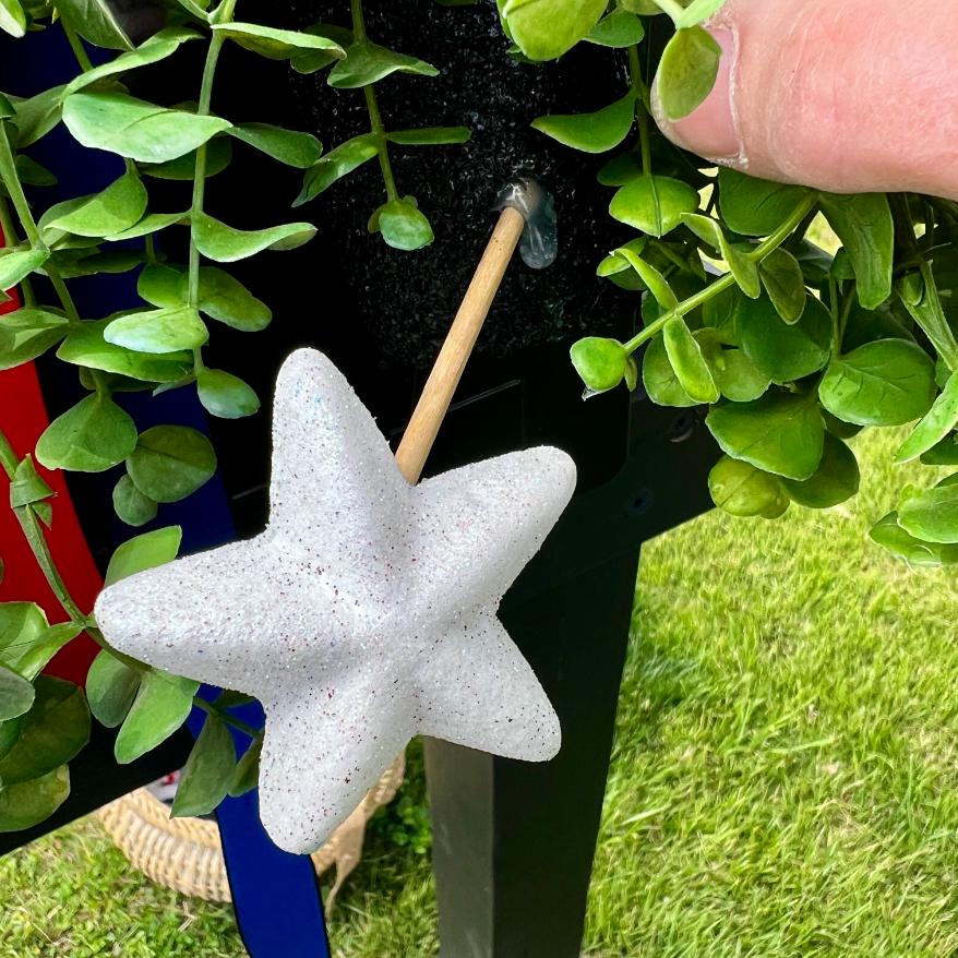 A hand holds a small rod with a white star-shaped object, inserting it into a hole in a black surface surrounded by green leafy plants—a perfect touch for your DIY patriotic mailbox swag. Grass and a woven basket are visible in the background.