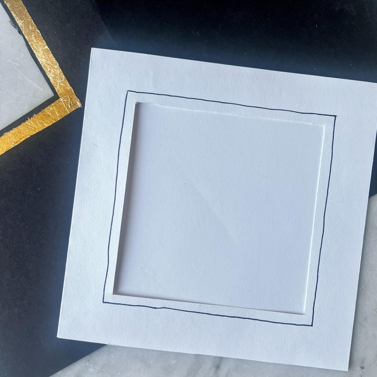 A white square card with a hand-drawn black border sits on a dark surface, showcasing the elegance of handmade frame cards. Part of another card with a gold foil corner design is visible in the background, adding to the modern, minimalist feel.