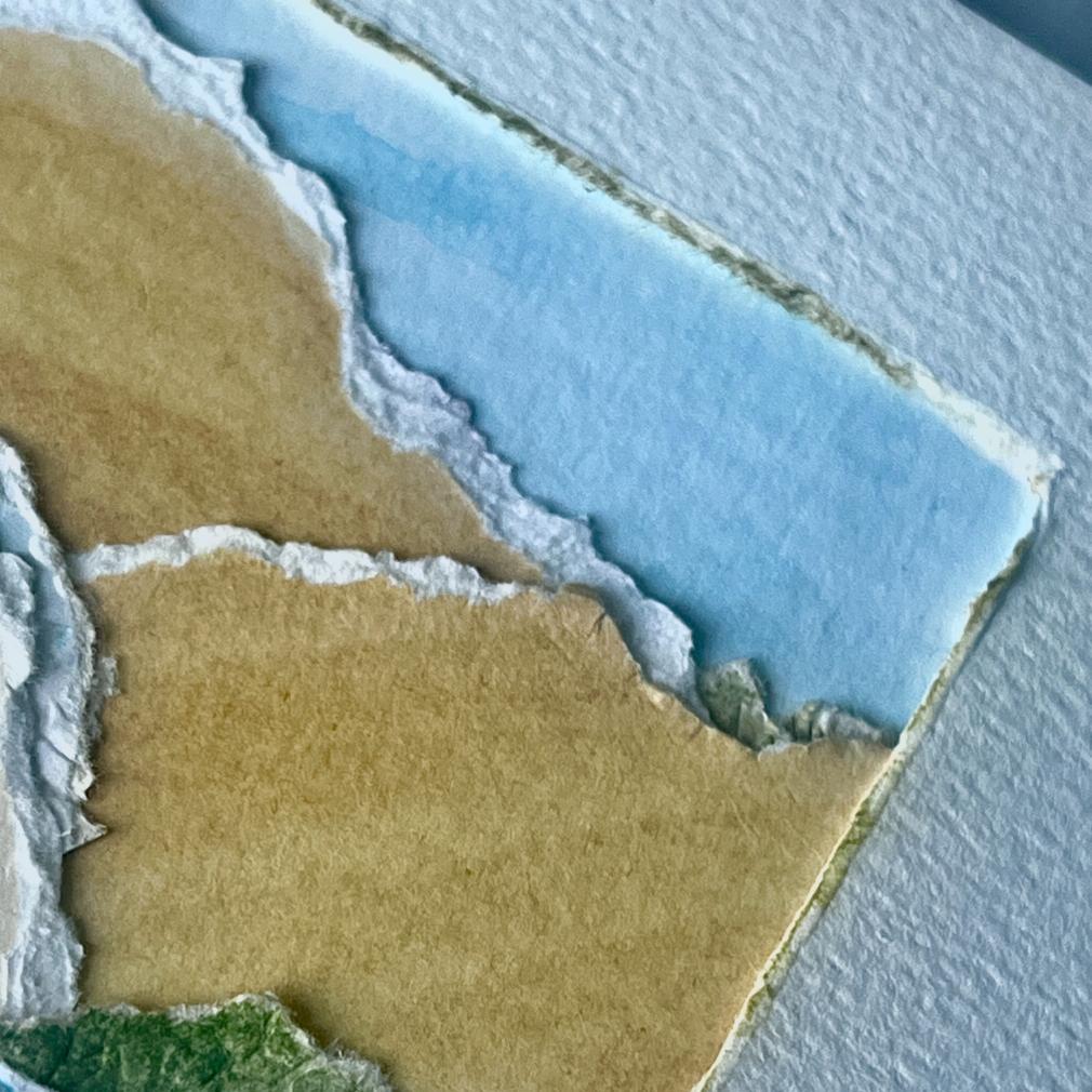 A close-up of layered, torn paper in shades of brown, white, green, and blue, arranged to resemble a textured landscape, possibly depicting land, grass, and sky on a textured white background.