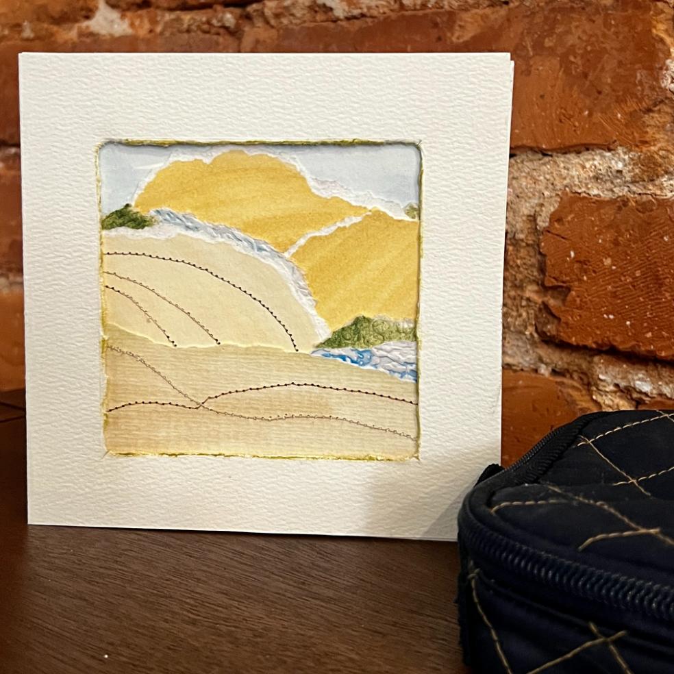 A textured card with an abstract landscape, showing yellow hills, green patches, a blue river, and stitched lines, stands against a red brick wall with a black quilted bag nearby.