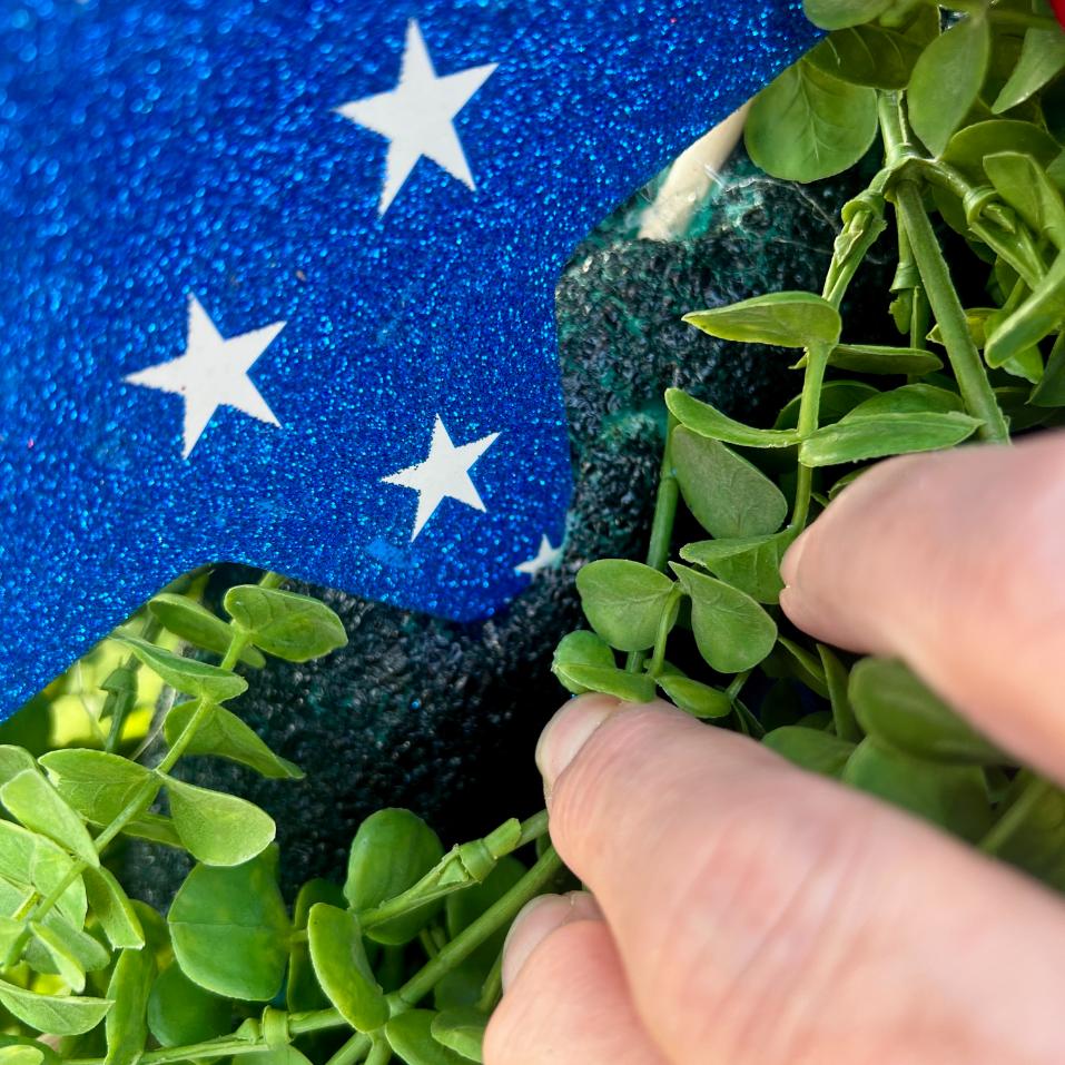 A close-up of a hand touching green leaves with a blue glittery object featuring white stars in the background, perfect for creating a DIY patriotic mailbox swag.