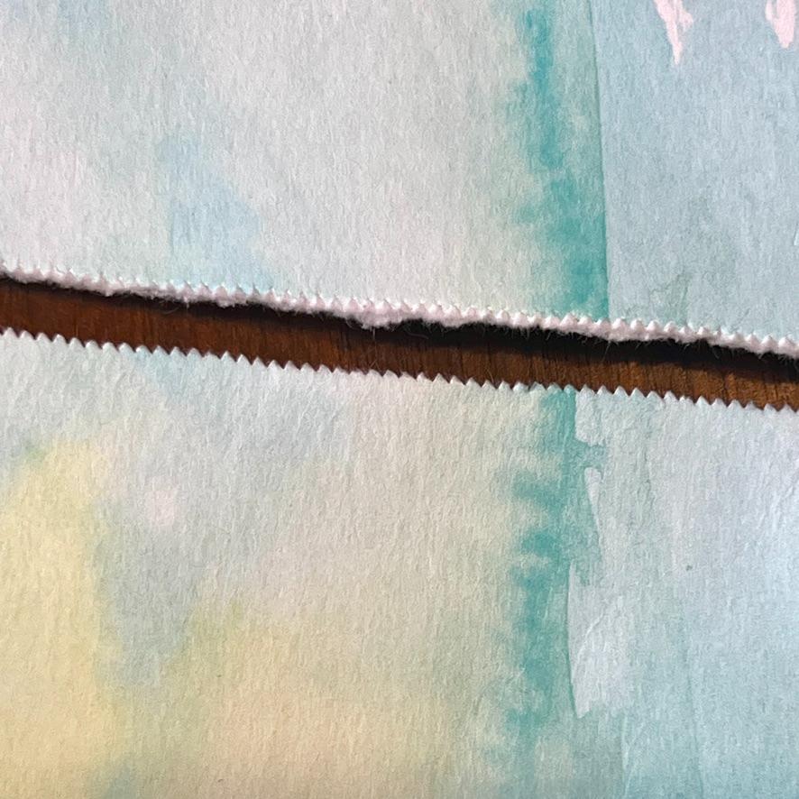A close-up of a piece of watercolor paper torn in a jagged line, revealing a textured brown surface beneath. The paper has a pale blue and yellow wash with subtle watercolor gradients.