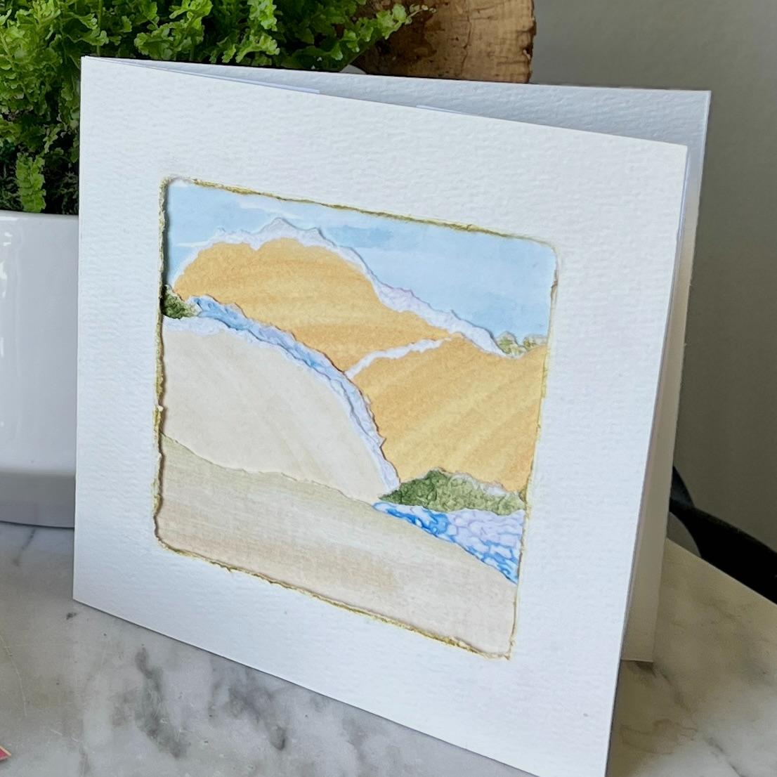 A handmade greeting card displayed upright shows a watercolor landscape with hills, a blue stream, and greenery, bordered by a textured square on white paper. A green potted plant is in the background.