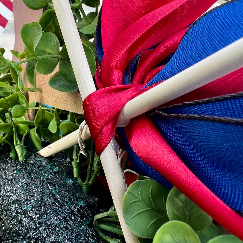 Close-up of DIY patriotic mailbox swag: red and blue fabric tied around wooden poles, surrounded by green leafy plants and a patch of dark artificial grass. An American flag and brown cardboard are partially visible in the background.