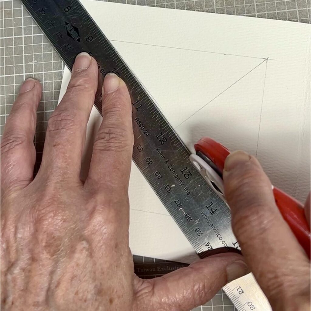 A person uses a metal ruler and a red utility knife to cut a piece of white textured paper along a penciled line on a gridded cutting mat.