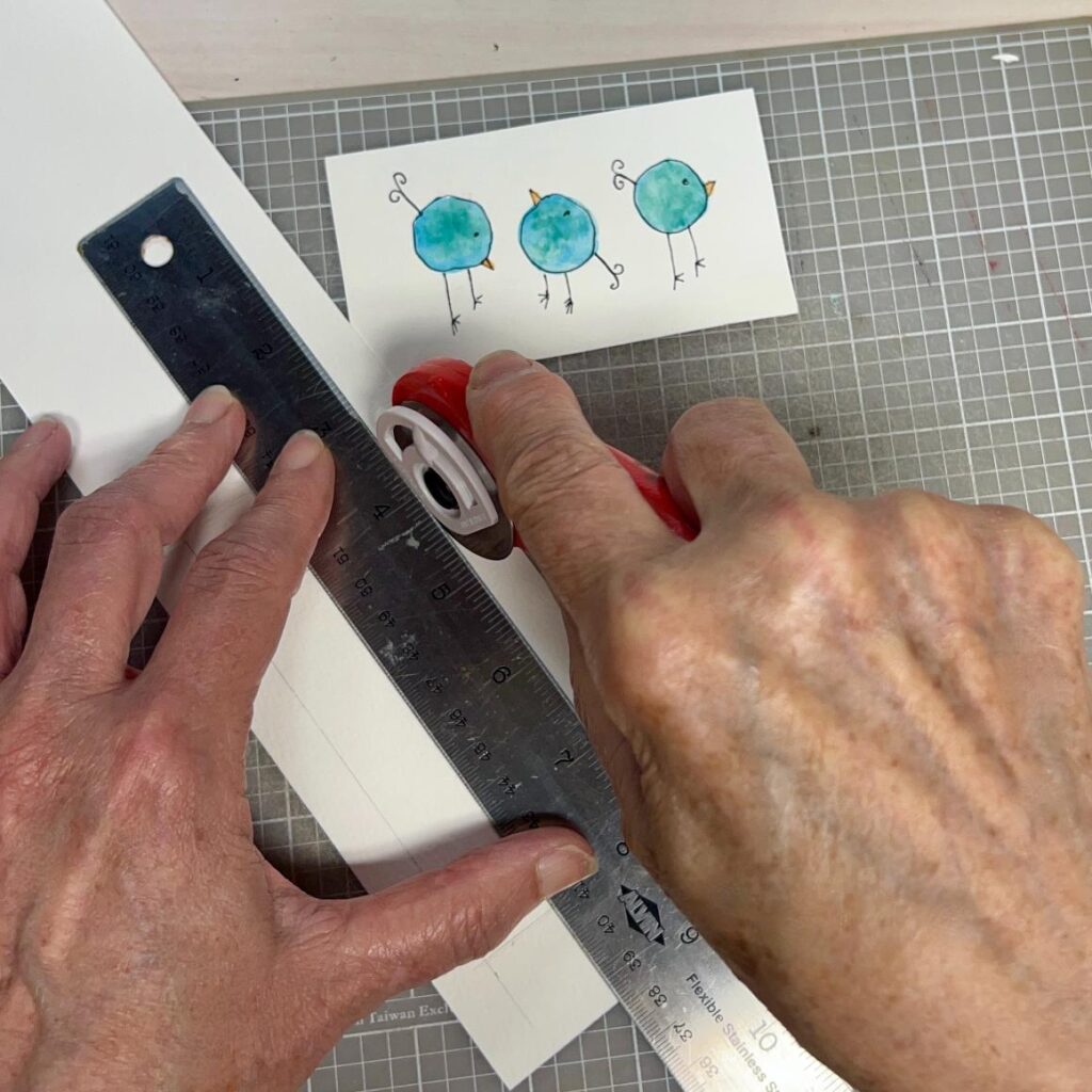 A person uses a rotary cutter and metal ruler to cut white paper on a grid mat. Above the hands is a card with three blue, round birds drawn and painted in a whimsical style.