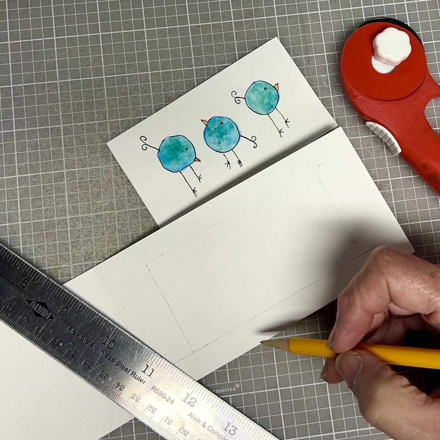 A hand uses a pencil and ruler to draw on white paper. Nearby are a red rotary cutter and a card featuring three simple, blue cartoon birds with orange beaks, all on a grid cutting mat.