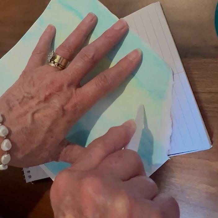 A person’s hands are shown using a bone folder to emboss a wavy line in a piece of paper with blue watercolor against a stack of lined index cards on a wooden surface.