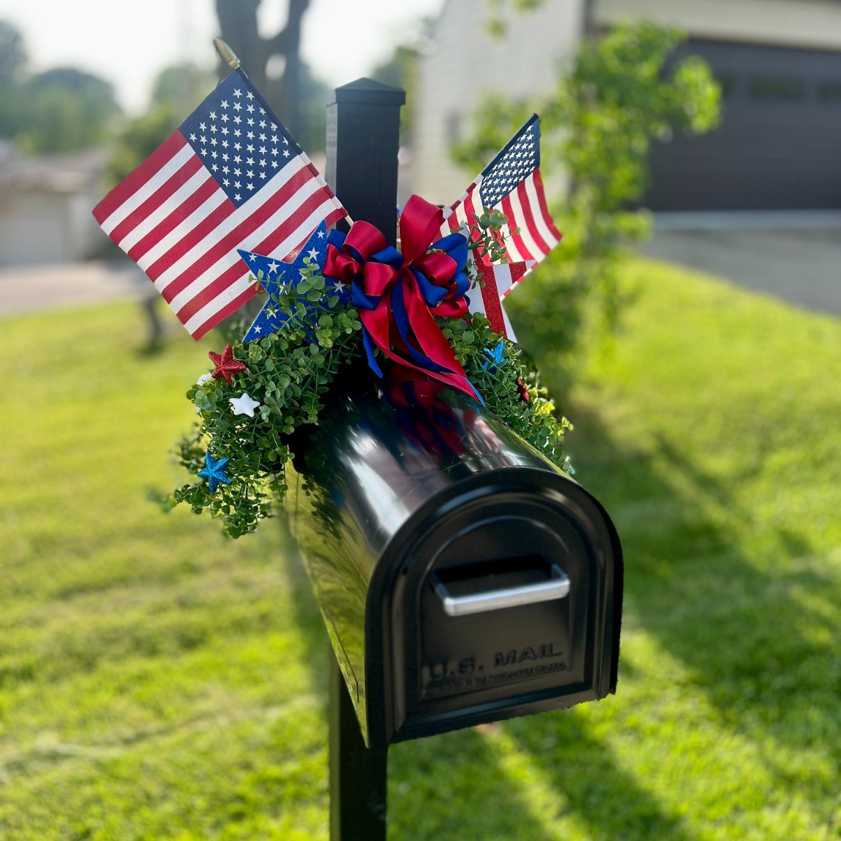 A black mailbox decorated with a DIY patriotic mailbox swag—featuring two American flags, red, white, and blue ribbons, and green foliage—stands on a lawn in front of a house, celebrating a patriotic occasion.