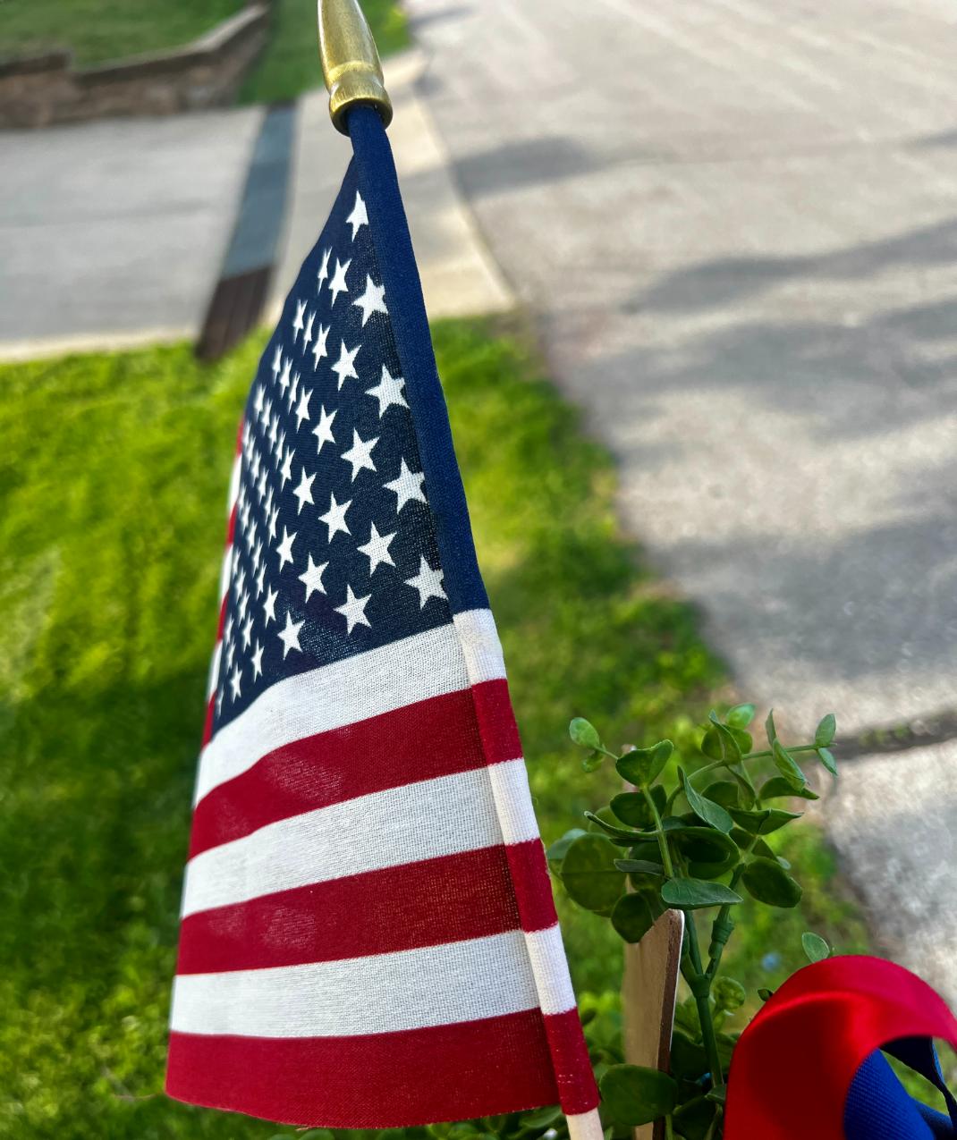 A small American flag on a stick is displayed outdoors next to greenery and a red ribbon, creating a festive DIY patriotic mailbox swag, with a sidewalk and street visible in the background.
