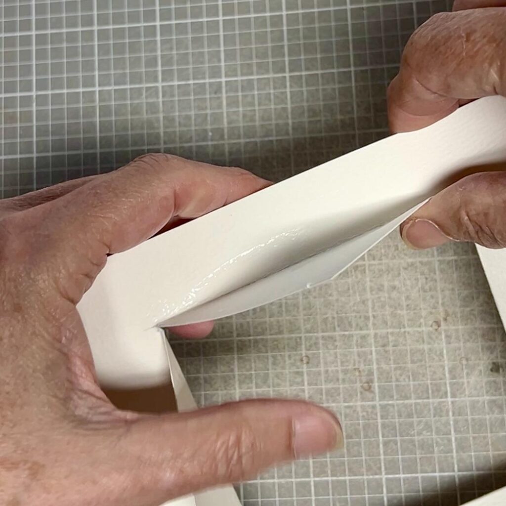 Close-up of two hands gluing and pressing together the edges of thick white paper, with a grid-patterned cutting mat underneath.
