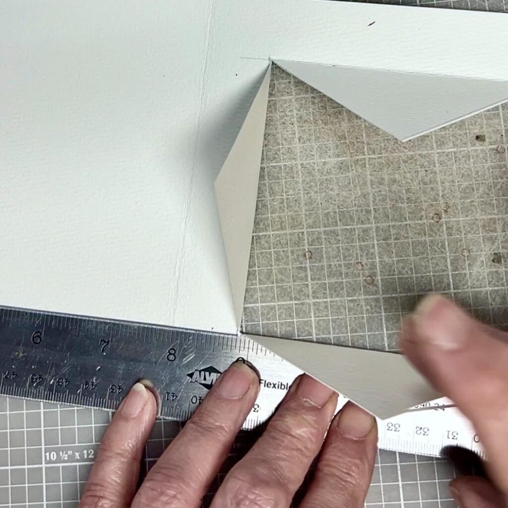 A close-up of hands folding the corner of thick white paper over a grid-patterned surface, using a metal ruler for precision. The person appears to be doing a craft or bookbinding project.