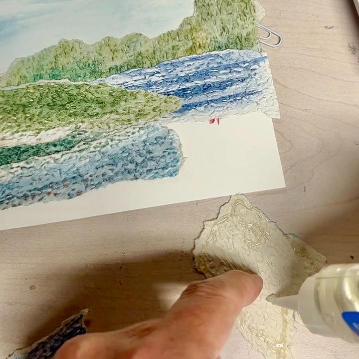 A hand applies glue to a textured piece of paper, preparing to attach it to a mixed media landscape artwork featuring green trees, blue water, and sky on a wooden surface.