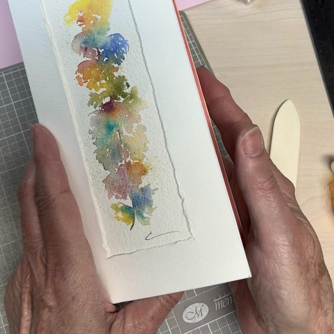 A pair of hands holds a card featuring an abstract, vertical watercolor painting in vibrant colors. The card has textured edges and is shown on a crafting table with a bone folder and other tools nearby.