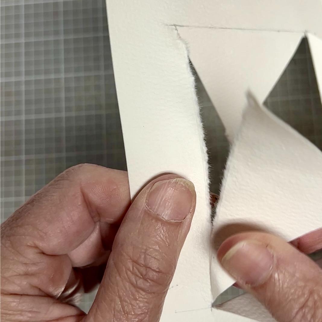 A close-up of hands tearing a piece of thick white paper along a cut line, creating a jagged opening shaped like a star on a grid-patterned surface.