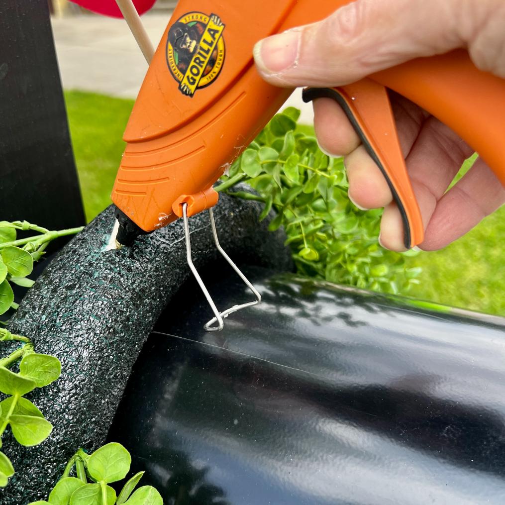 A hand uses an orange Gorilla hot glue gun to apply glue to a black foam ring for a DIY patriotic mailbox swag, next to green artificial leaves, outdoors on a sunny day.