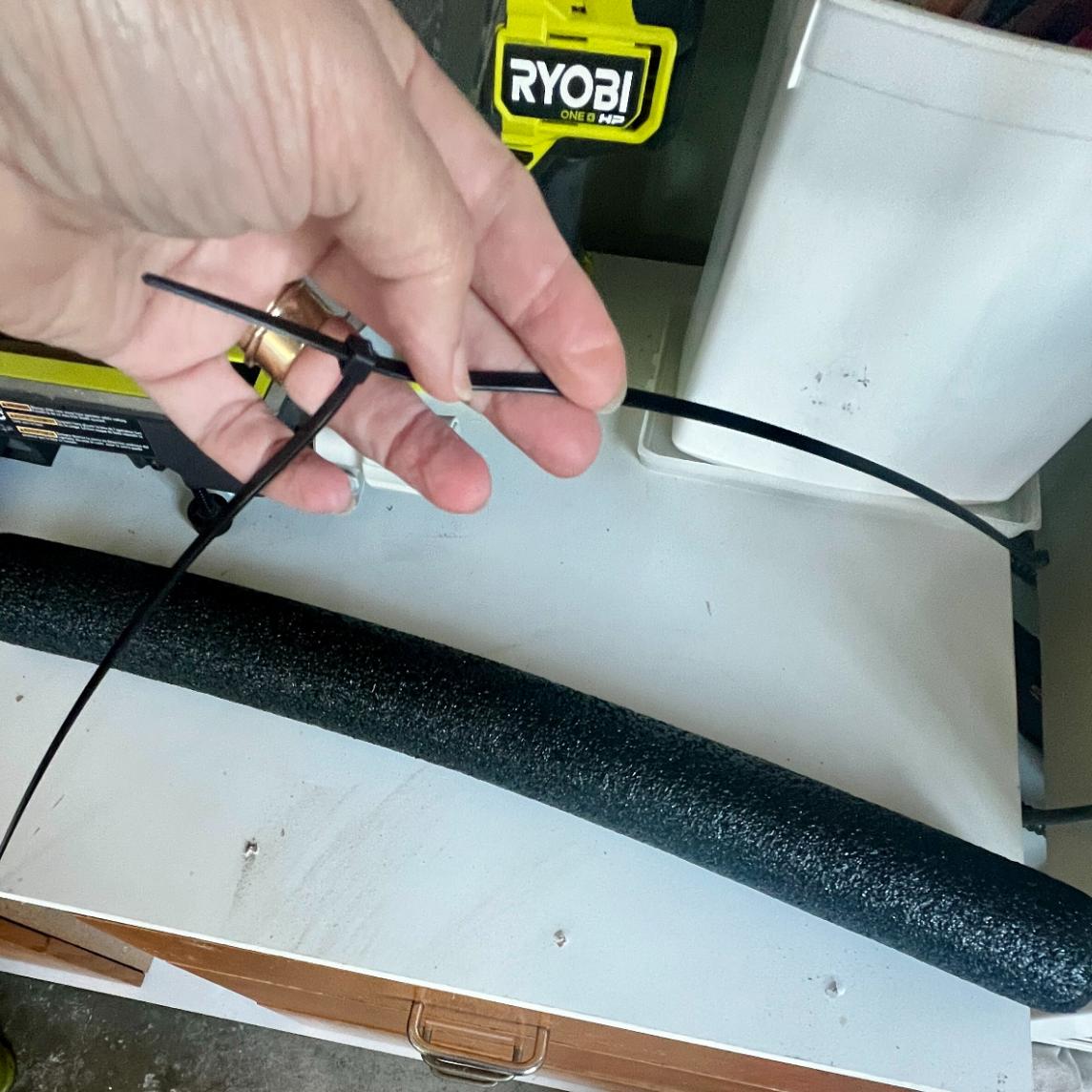 A hand tightens a black zip tie around a foam pipe cover, with tools—including a Ryobi tool—scattered on a white workbench in the background, ready for assembling DIY patriotic mailbox swag.