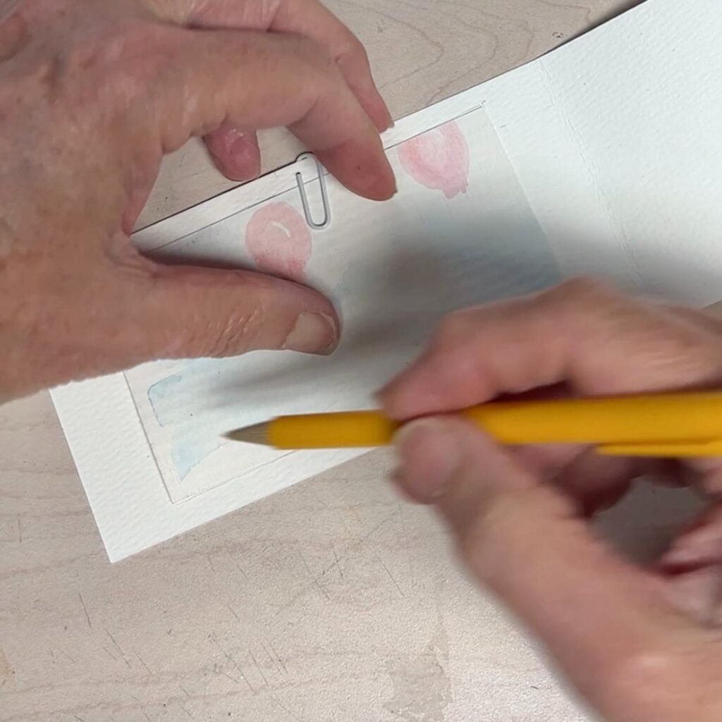 A person uses a yellow pencil to draw on a small piece of watercolor paper, which is held in place on a white folder with a paperclip.