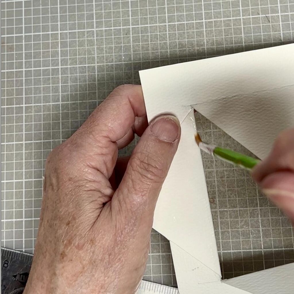 A person uses a small brush to apply glue or adhesive to the edge of a cut-out piece of white paper on a grid-patterned cutting mat. A metal ruler is visible in the corner.