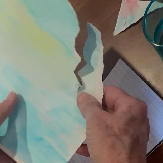 Two hands are tearing a piece of paper painted with light blue and yellow watercolor. The paper is held over a lined notepad on a wooden table, with a glass and another torn paper visible nearby.