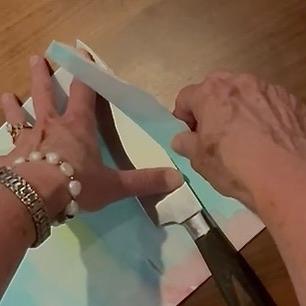 A person wearing a bracelet and ring uses the side of a knife to tear a piece of pastel-colored paper.