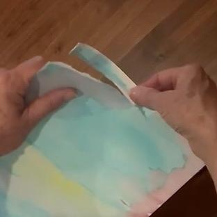 Hands tearing a piece of pastel-colored paper, with a light blue and yellow wash, over a wooden surface.