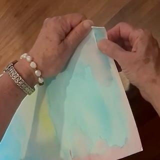 A person wearing a bracelet and watch carefully peels back the corner of a sheet of watercolor paper painted in soft blue and pastel tones.