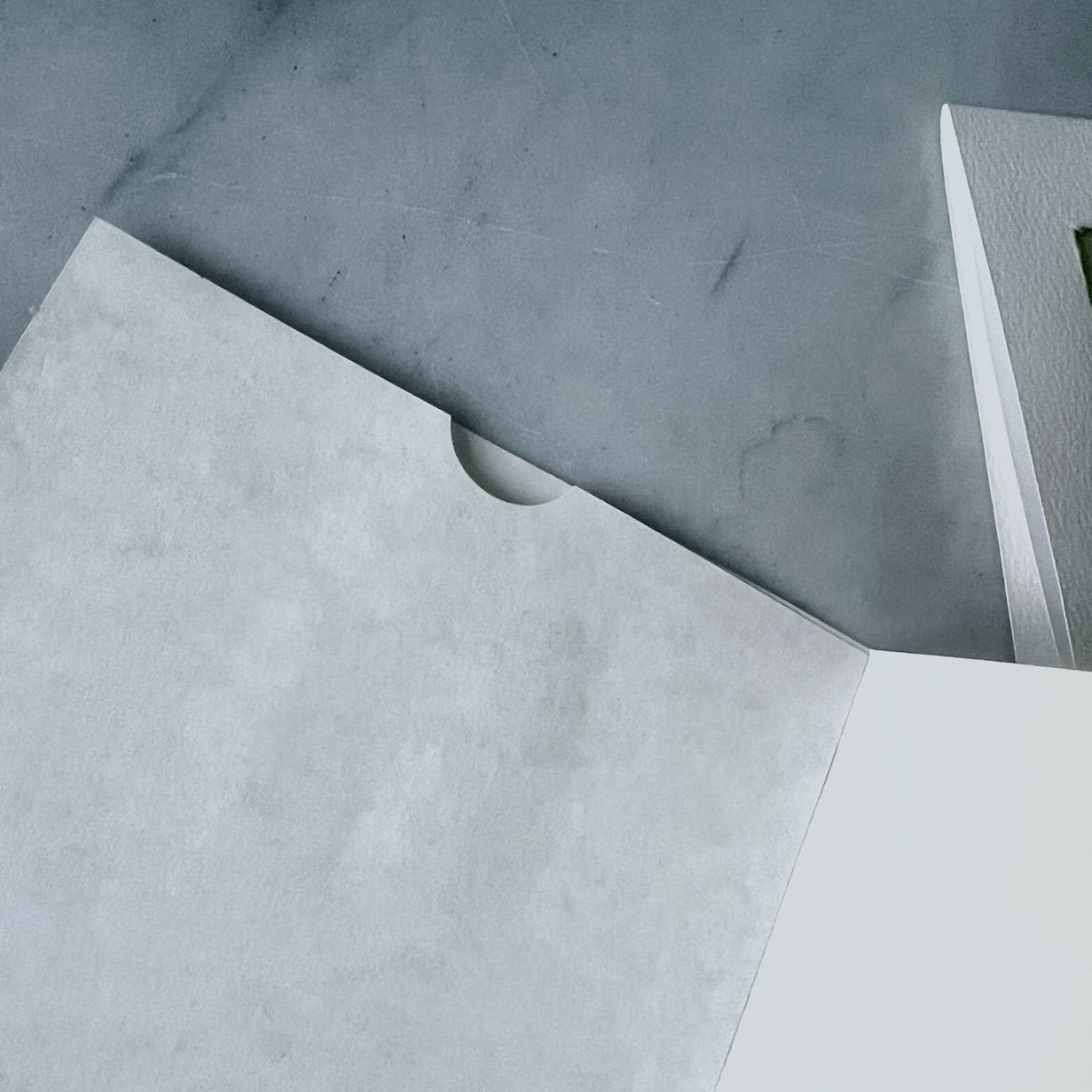 A close-up of a minimalist grey folder with a semicircular cutout, partially open on a grey surface next to a white rectangular object.