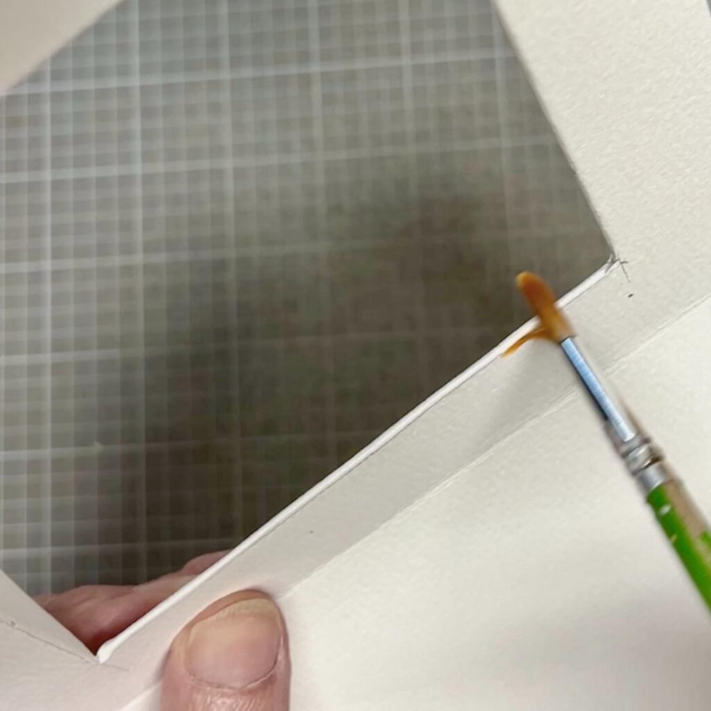 A hand holds a white mat board with a rectangular cut-out while using a small paintbrush to apply glue or adhesive along the inner edge. A grid-patterned surface is visible in the background.