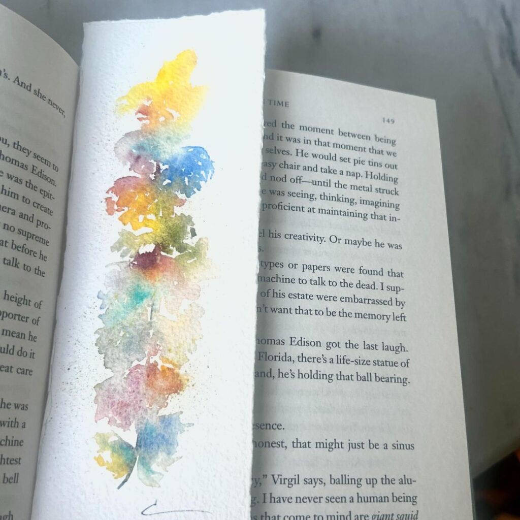 A watercolor bookmark with abstract, colorful leaf shapes rests on the open pages of a book. The background surface is light gray.