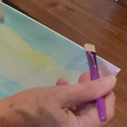 A hand holds a flat purple paintbrush above a piece of watercolor paper with light blue and yellow paint washes, on a wooden surface.