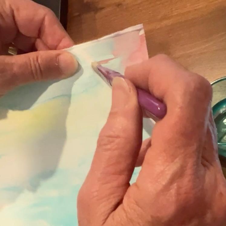 A person uses a paintbrush to apply water on a piece of paper, with their hands visible and a wooden surface in the background.